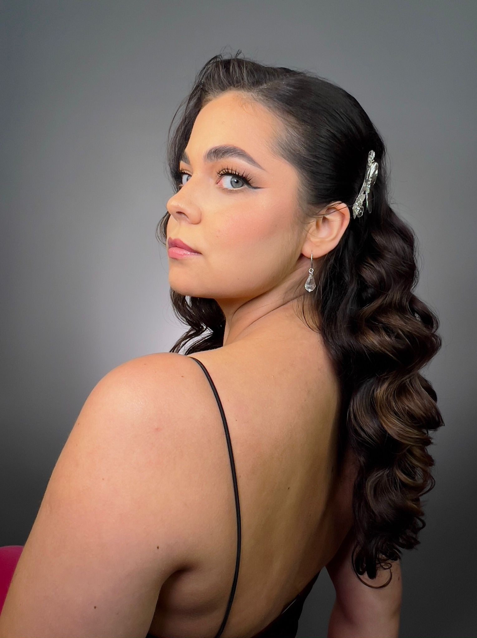 Kobieta z profesjonalnym makijażem oczu i upiętymi, pofalowanymi włosami, ozdobionymi srebrną spinką i kolczykami, spogląda przez ramię na szarym tle.