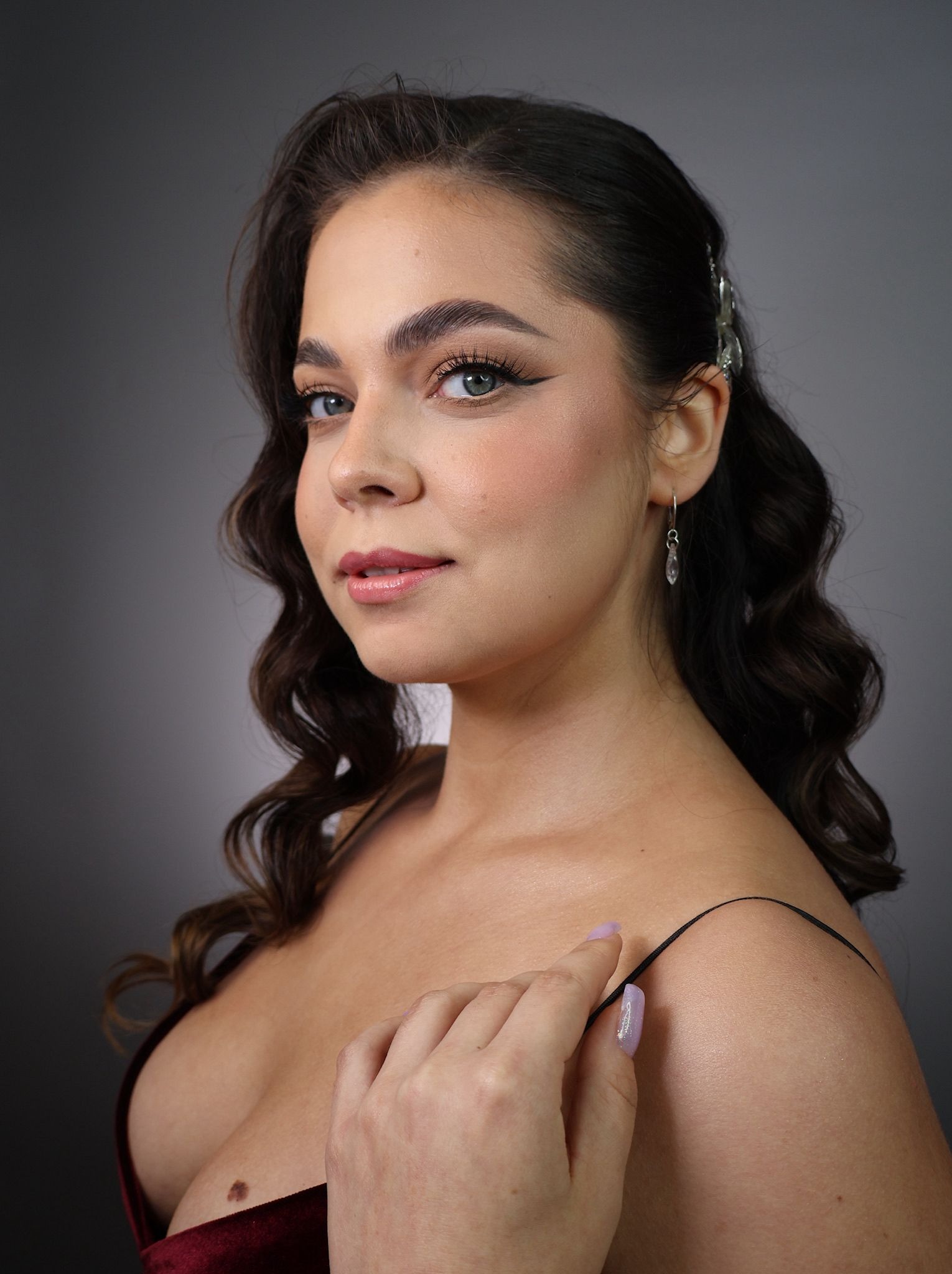 Portret kobiety z profesjonalnym makijażem: wyraźna kreska na powiece, delikatny róż na policzkach, błyszczące usta. Upięte włosy z ozdobną spinką i subtelne kolczyki.