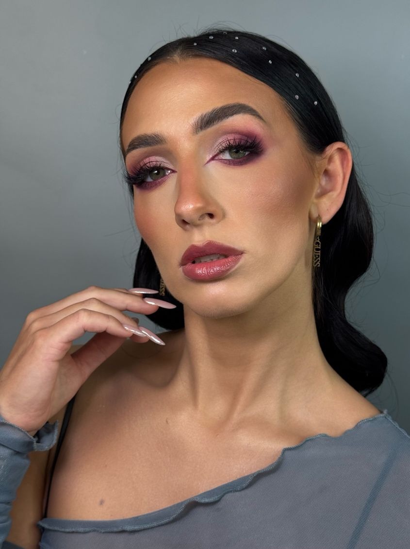 Portret kobiety z intensywnym makijażem oczu w odcieniach różu i fioletu, długimi paznokciami i kolczykami Guess na szarym tle.