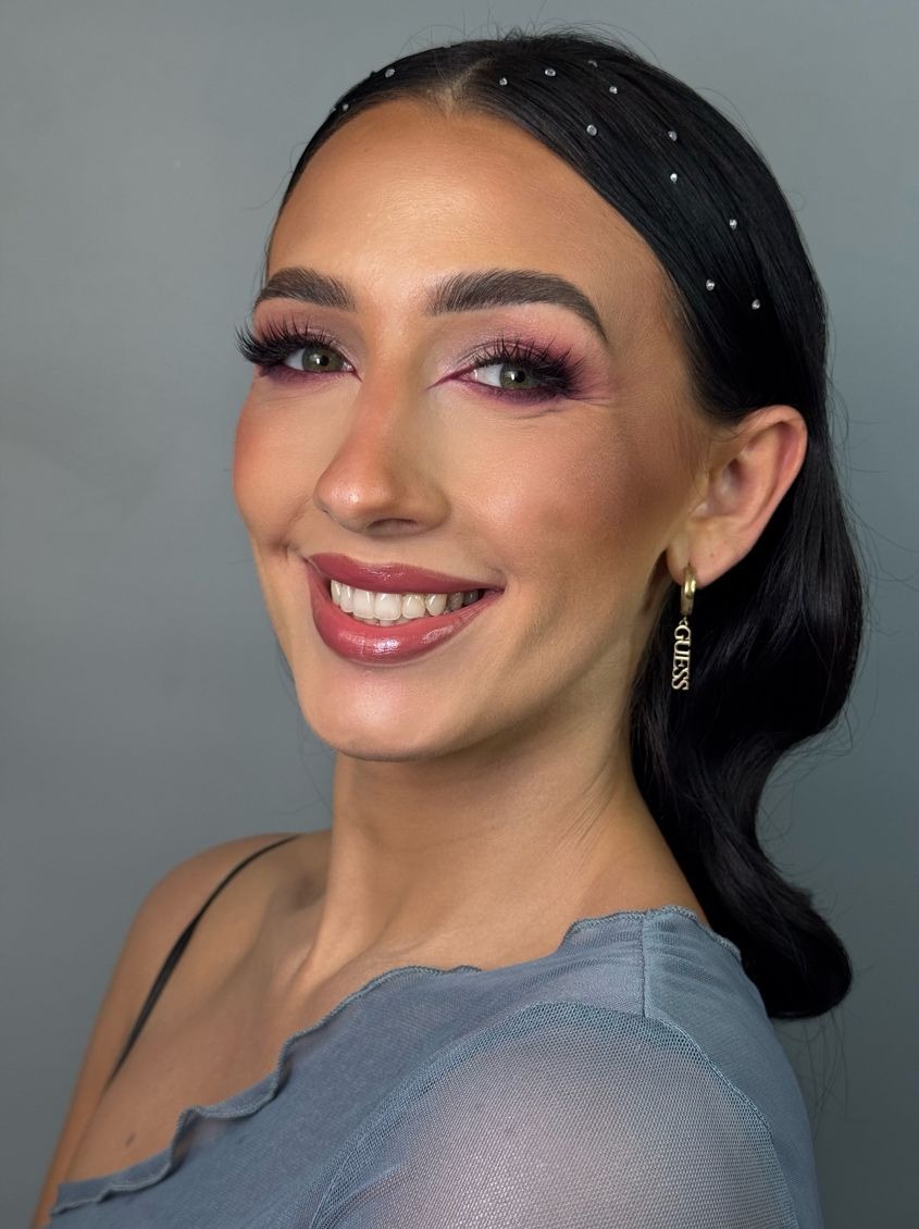 Uśmiechnięta brunetka z wieczorowym makijażem oczu w odcieniach różu i błyszczącymi ustami, ozdobną opaską i kolczykami Guess na szarym tle.