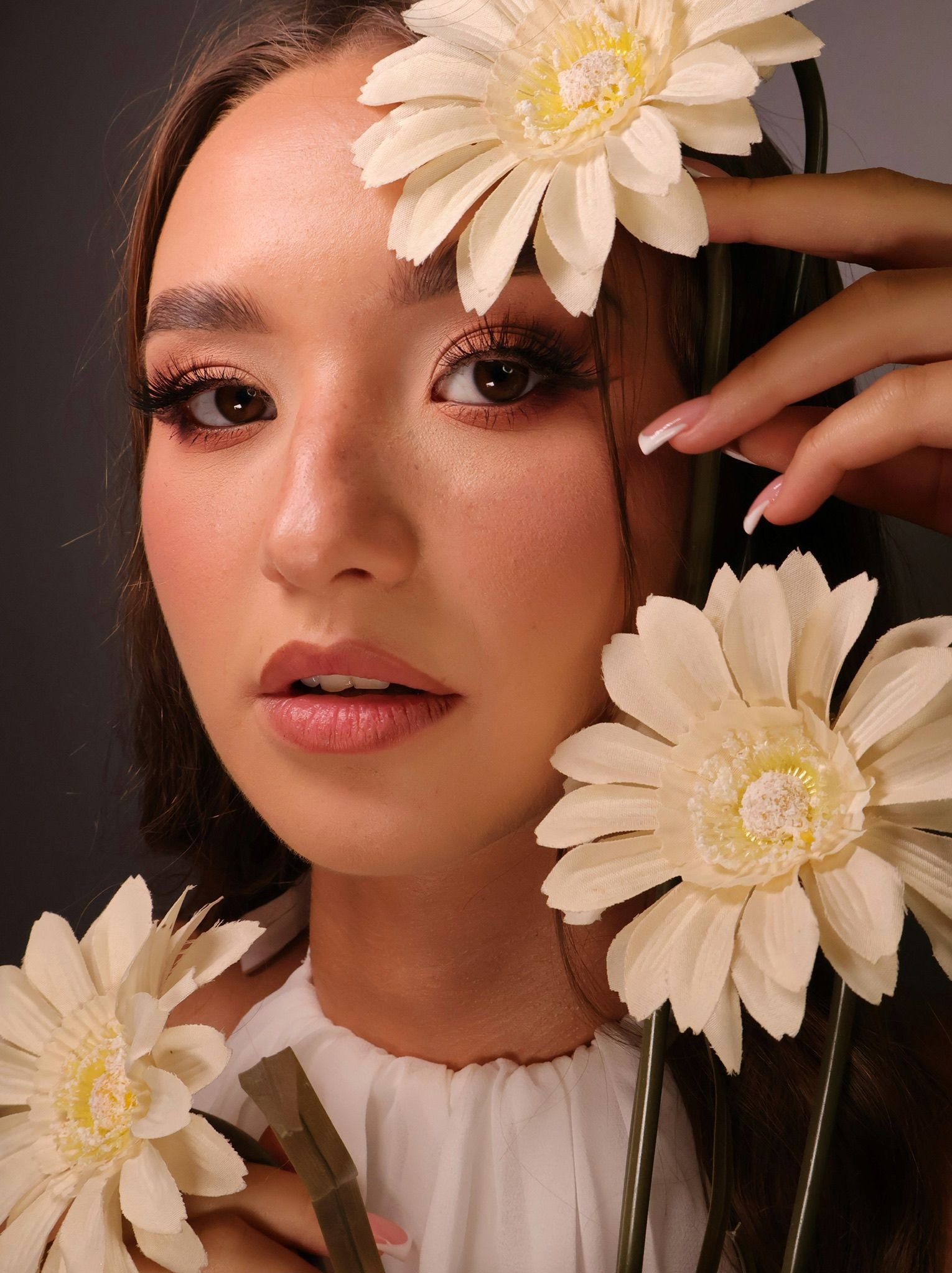 Portret kobiety z delikatnym makijażem, ozdobionej białymi kwiatami i z francuskim manicure, na ciemnym tle. Uwagę przyciągają długie rzęsy i subtelny błysk na ustach.