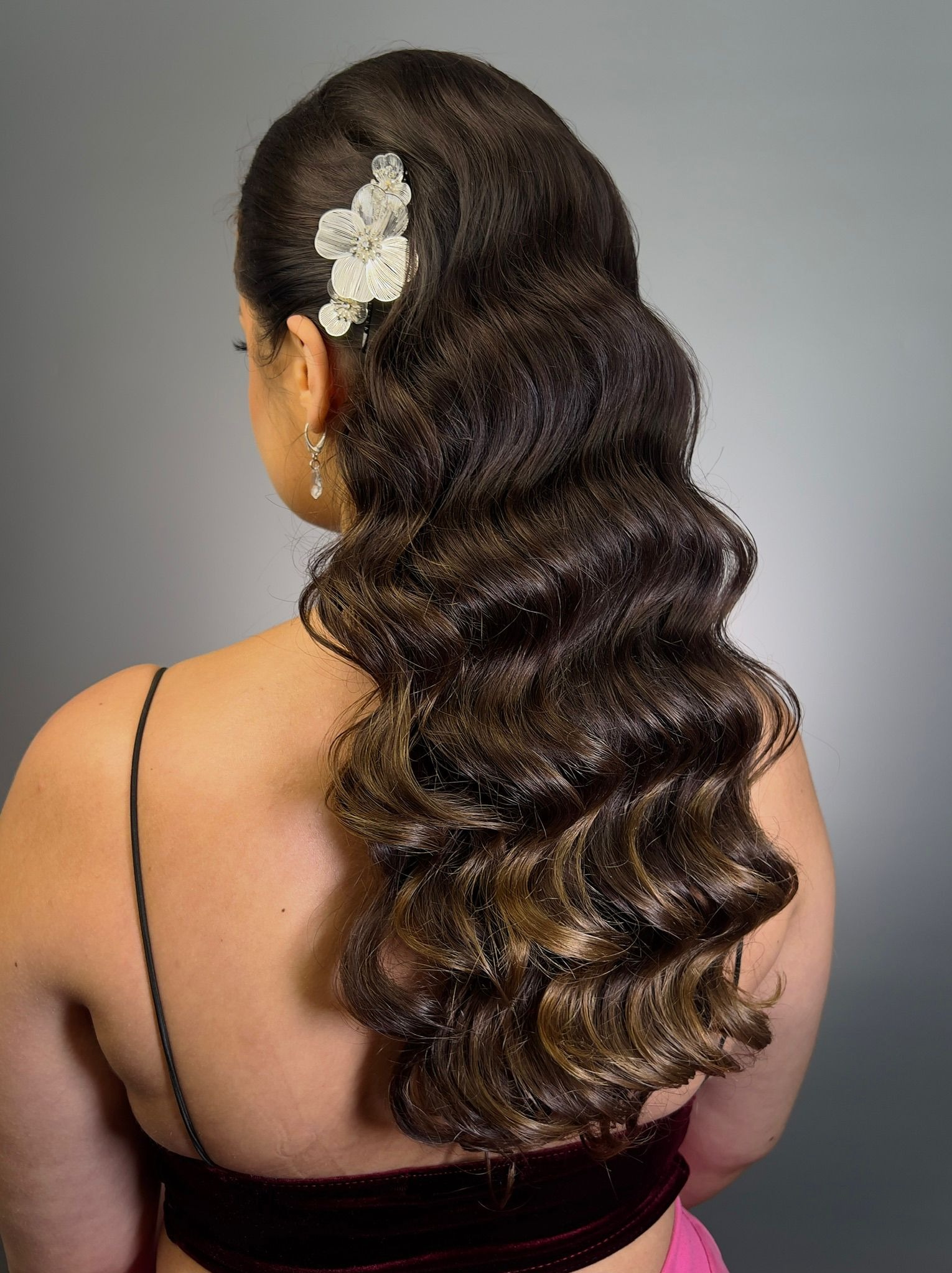 Upięcie z loków ombre, ozdobione kwiatową spinką i kolczykami, prezentowane z tyłu na tle szarego gradientu. Elegancka fryzura na specjalną okazję.