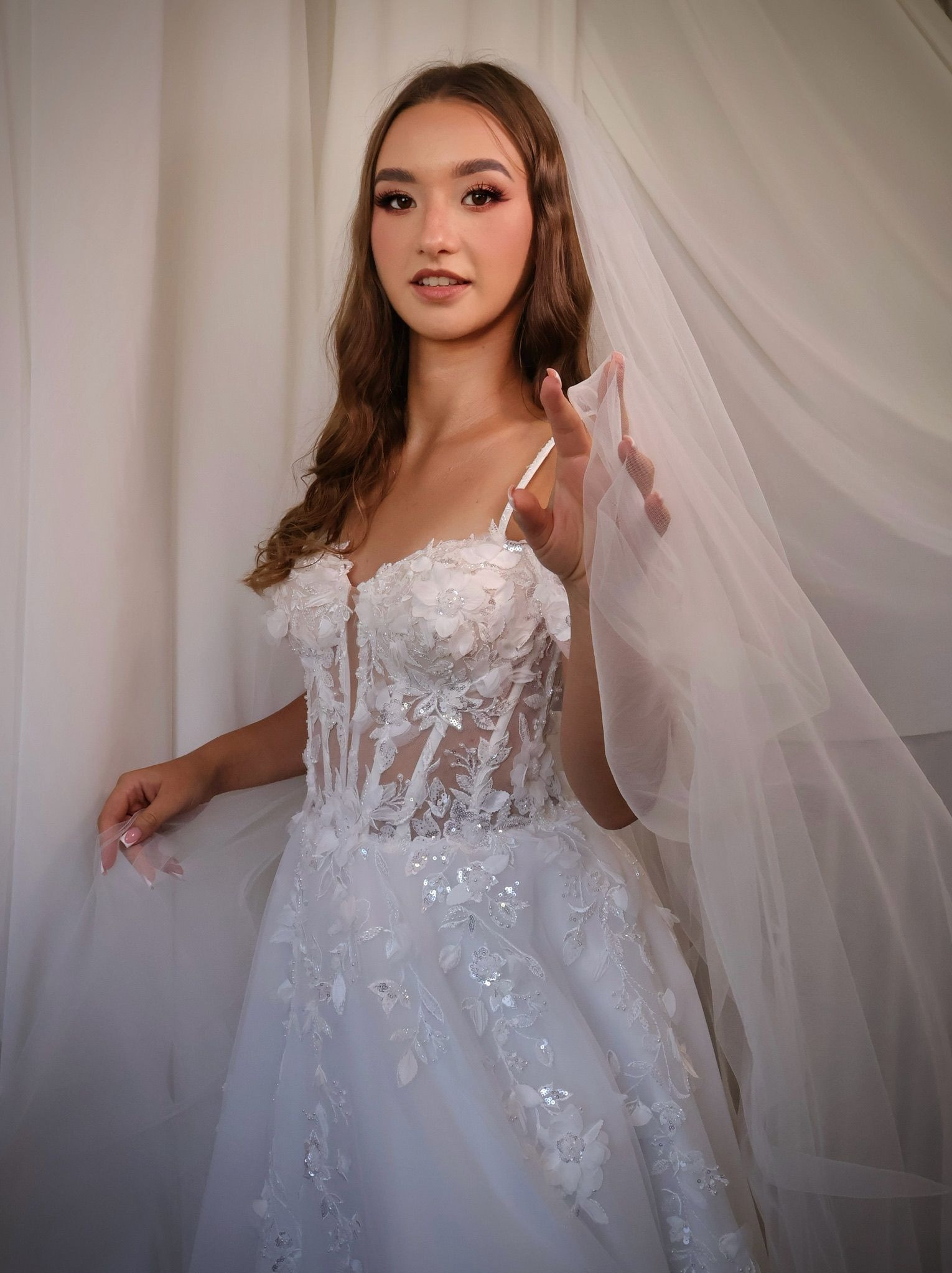 Panna młoda w koronkowej sukni ślubnej z welonem, delikatny makijaż oczu z różowym akcentem, naturalne fale. Ujęcie z bliska, jasne tło.