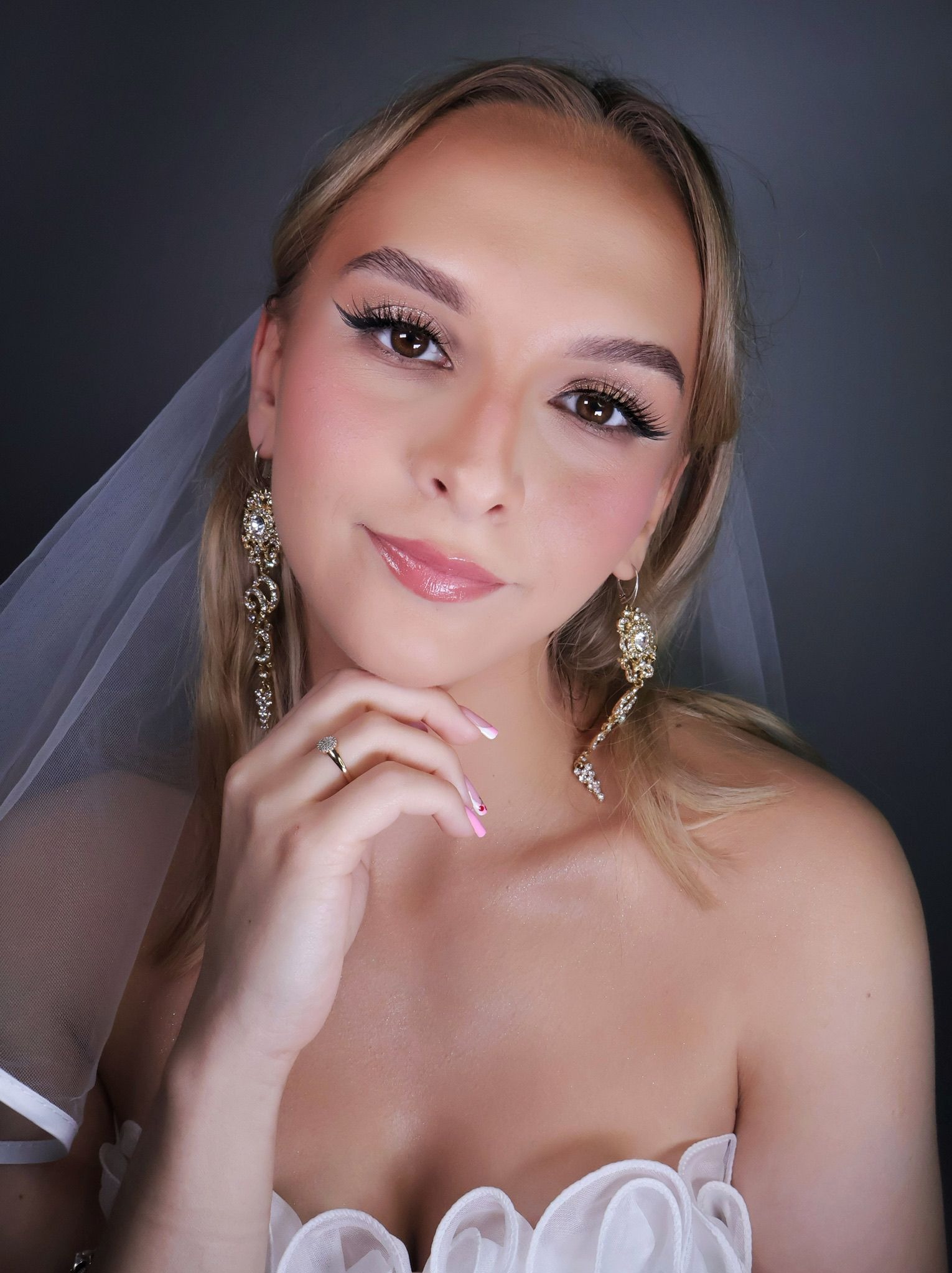 Panna młoda z delikatnym makijażem ślubnym, długimi rzęsami i błyszczącymi ustami. Widoczny manicure z różowymi akcentami i biżuteria ślubna. Welon w tle.