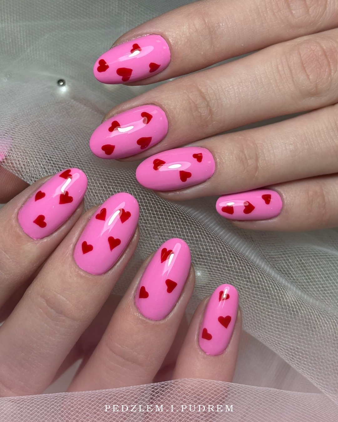 Różowe paznokcie w kształcie migdała z czerwonymi serduszkami, na tle delikatnej, białej tkaniny. Romantyczny, walentynkowy wzór na zadbanych dłoniach.