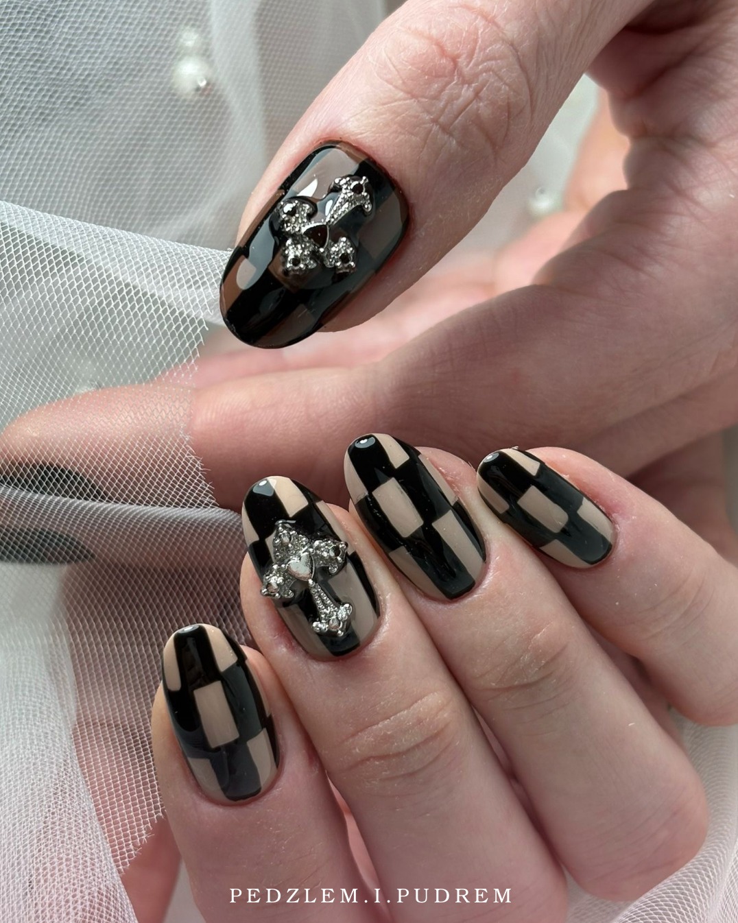 Dłoń z manicure w szachownicę beżowo-czarną, zdobiona srebrnymi krzyżami. Efektowny, błyszczący design na tle delikatnej, białej tkaniny.