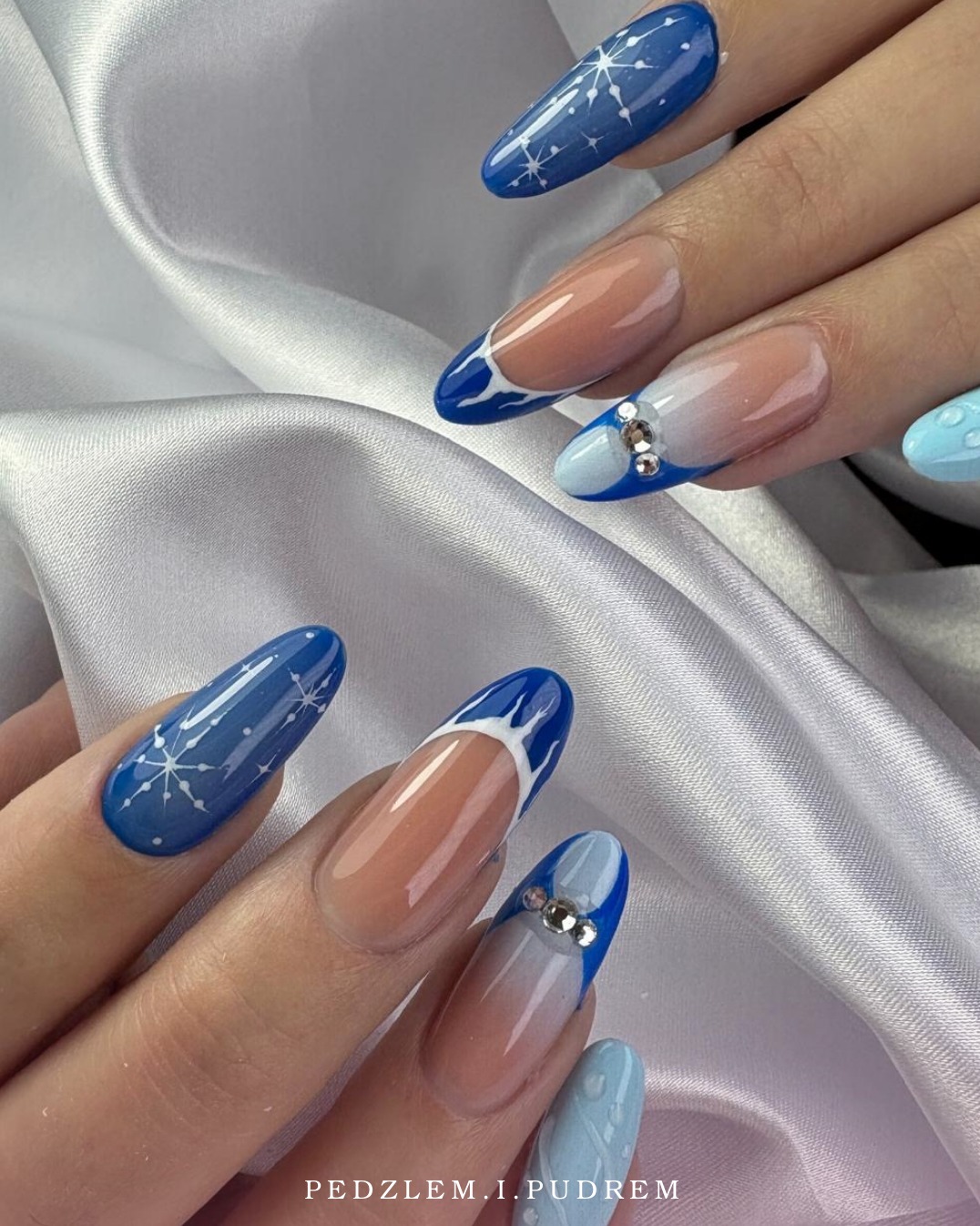 Zimowy manicure: niebieskie paznokcie z motywem śnieżynek, french z białym zdobieniem i cyrkonie na tle satynowej tkaniny. Elegancki i świąteczny.