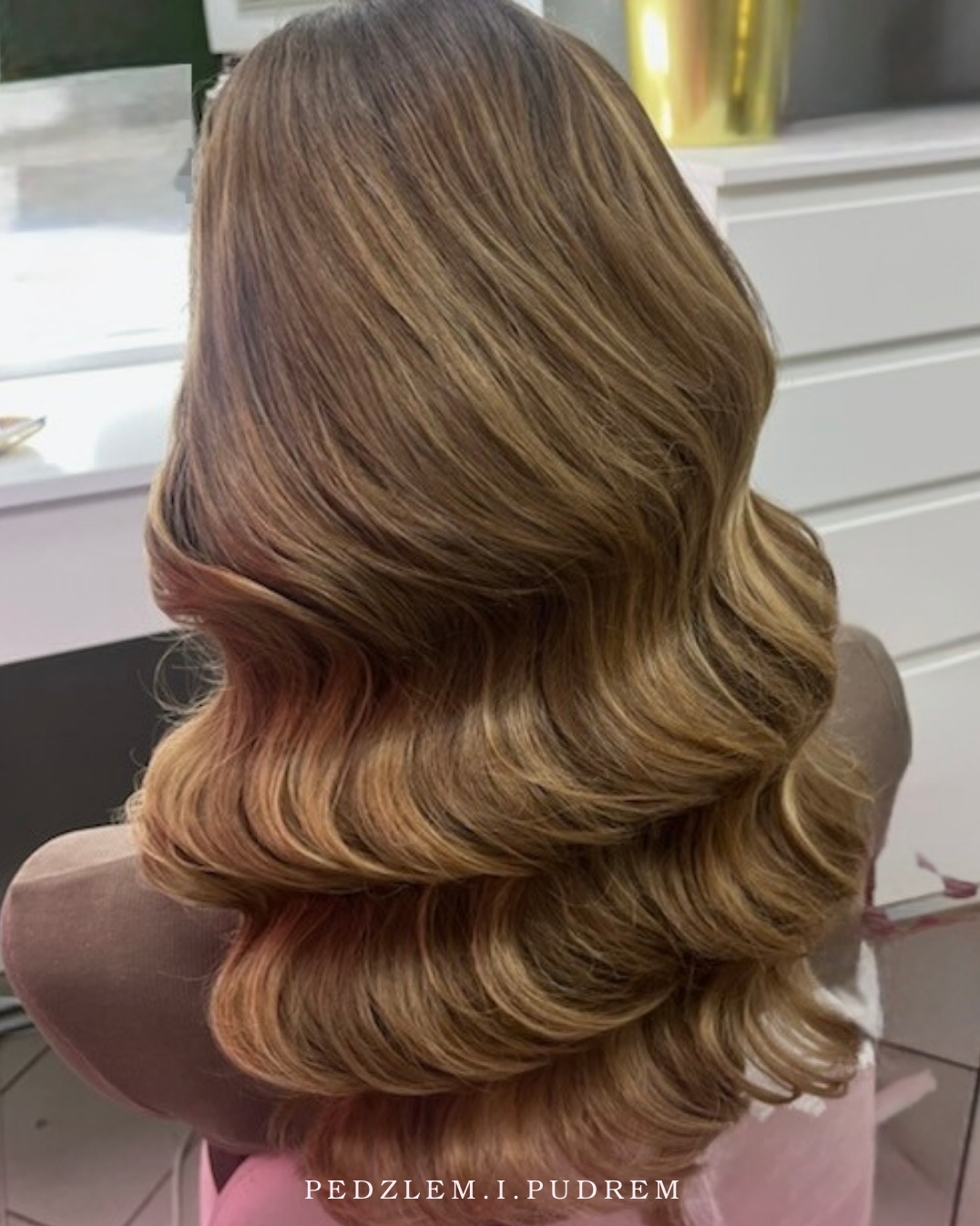 Kunsztownie ułożone, falowane włosy w odcieniach blondu i brązu, widok z tyłu, prezentujące efekt stylizacji na manekinie fryzjerskim w salonie.