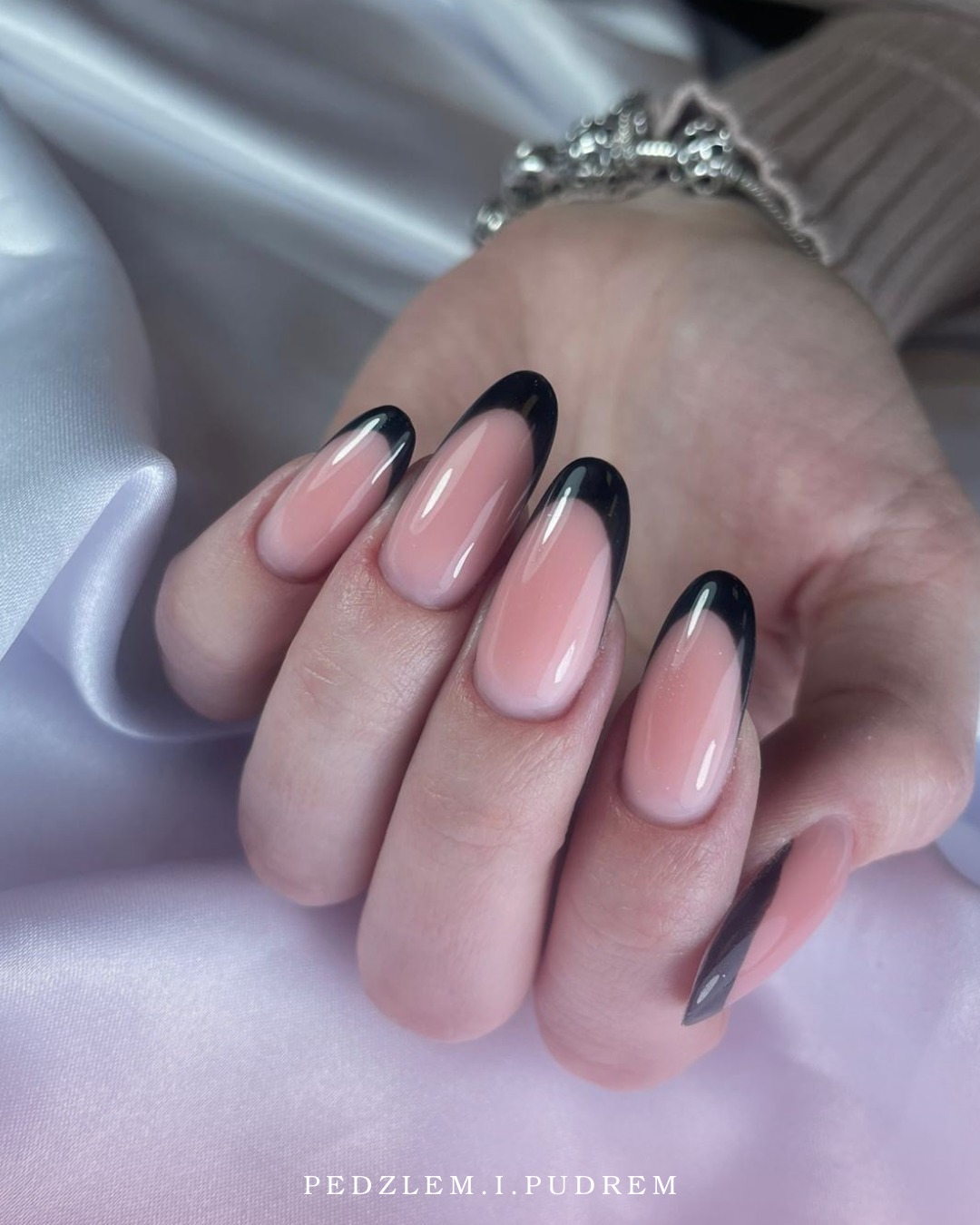 Eleganckie paznokcie z czarnym frenchem na tle satynowej tkaniny, ozdobione srebrną bransoletką. Długie, owalne paznokcie w odcieniach różu i czerni.