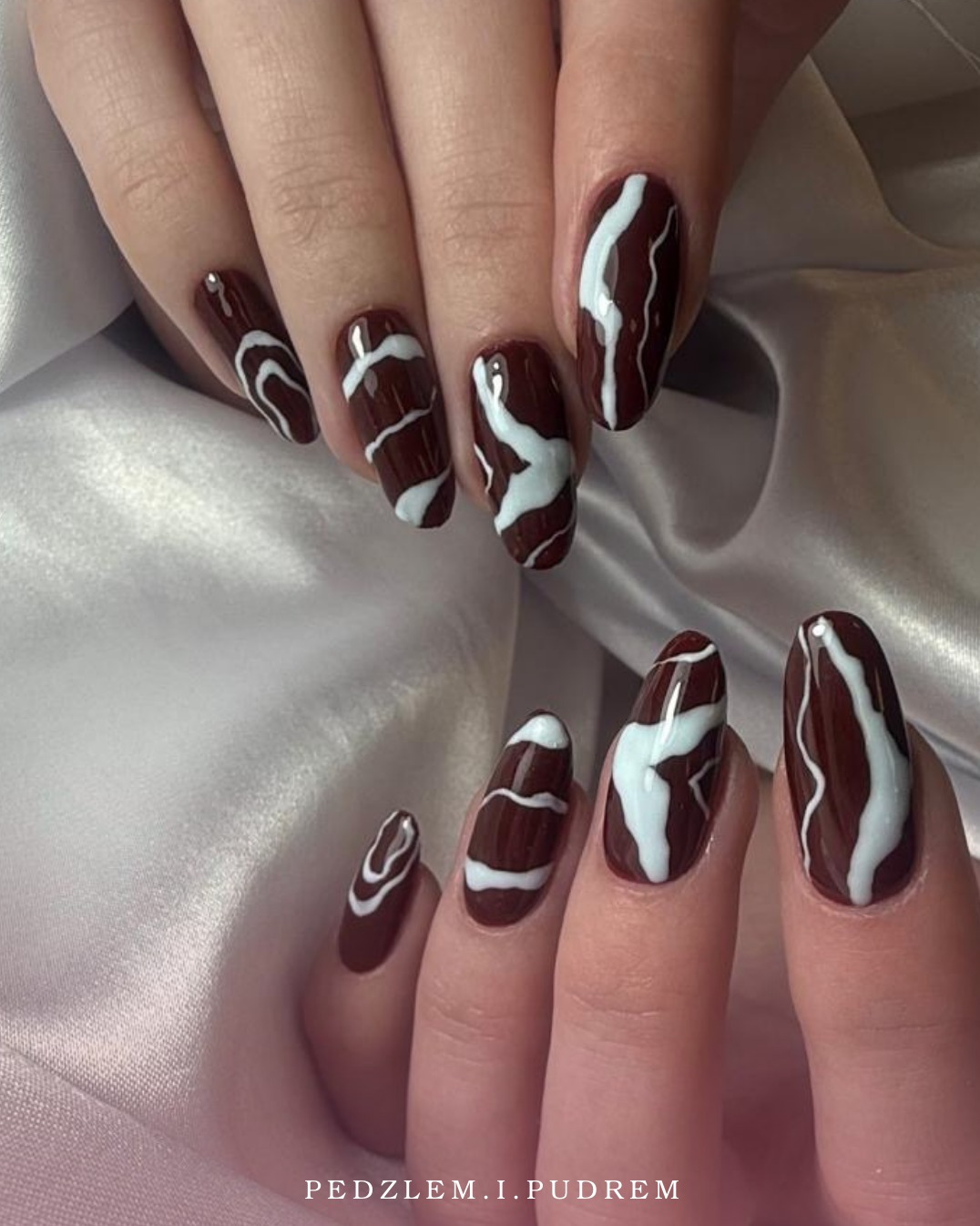 Dłonie z brązowymi paznokciami zdobionymi białymi, abstrakcyjnymi wzorami na satynowym tle. Widoczne logo 'PEDZLEM.I.PUDREM'.