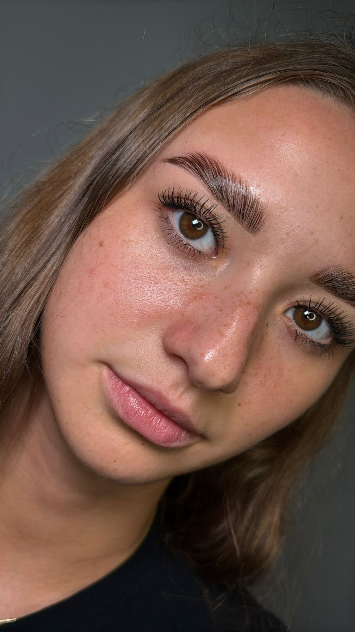 Portret kobiety z wyraźnie wystylizowanymi brwiami i podkreślonymi rzęsami. Widoczne piegi na twarzy i delikatny makijaż ust. Ujęcie z bliska, z boku.