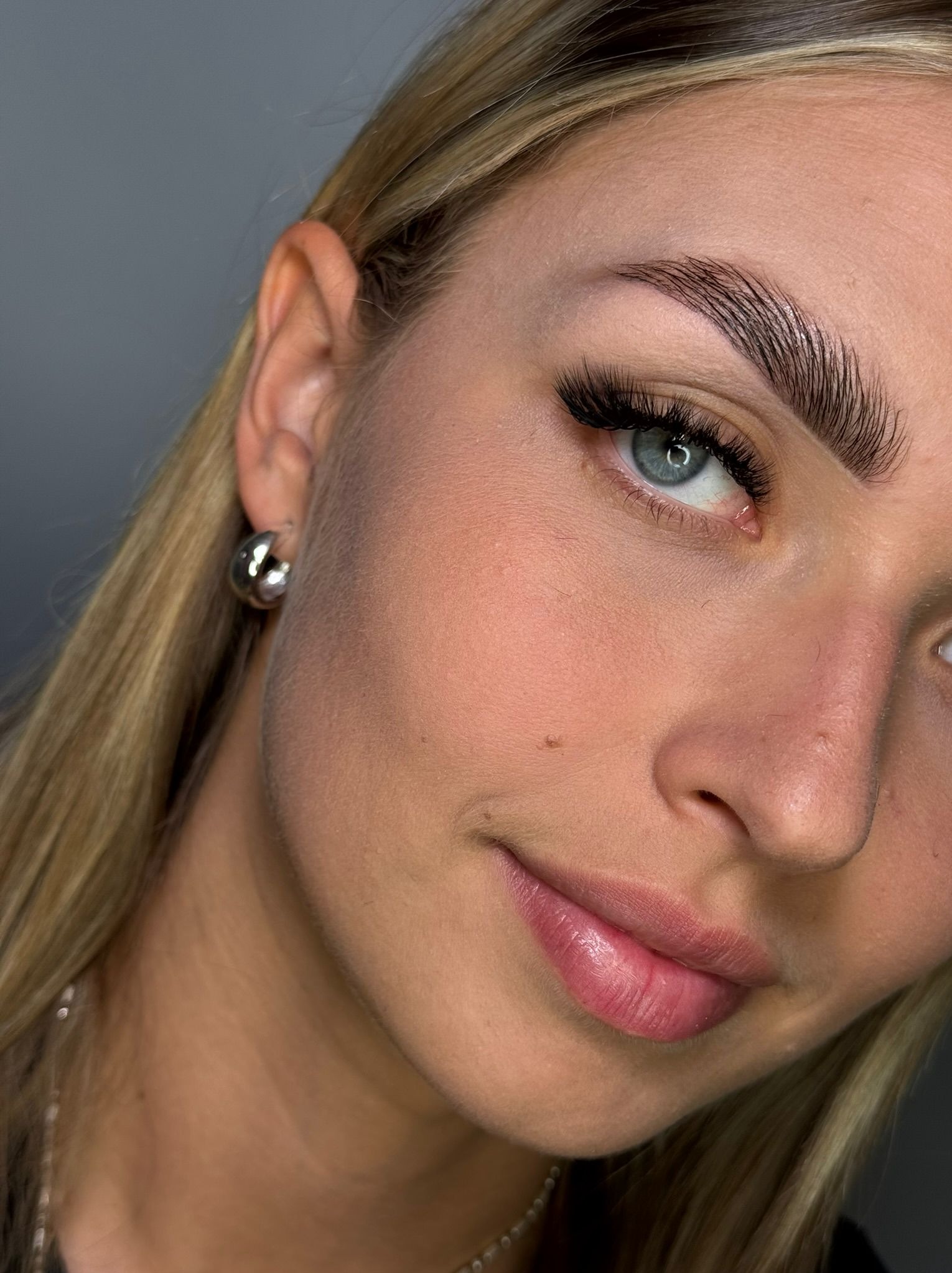 Zbliżenie na twarz kobiety z wyraźnie wystylizowanymi brwiami i sztucznymi rzęsami. Widoczny srebrny kolczyk w uchu i delikatny makijaż ust.