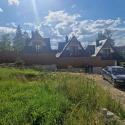 Trzy domy murowane z ciemnymi dachami i drewnianą elewacją, częściowo zasłonięte drewnianym ogrodzeniem, widoczne zza zarośniętego trawnika w słoneczny dzień.