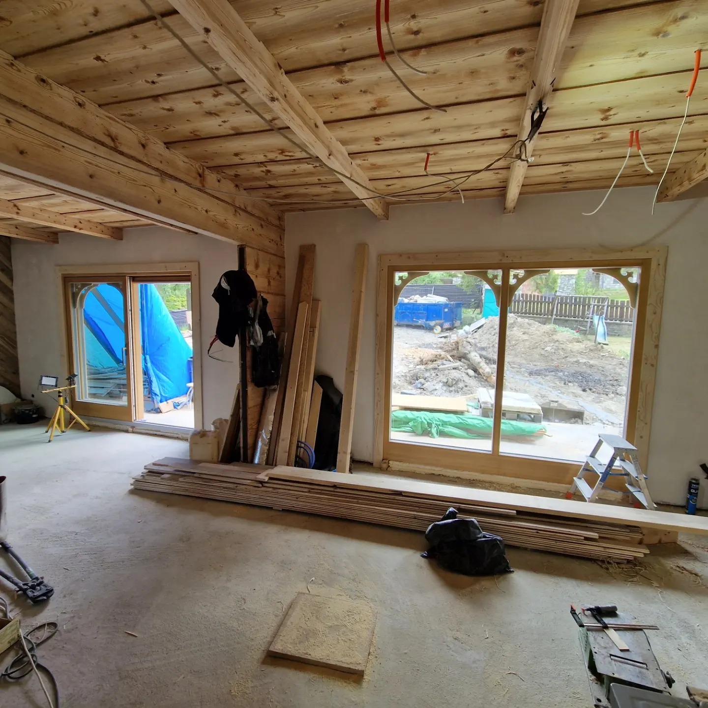 Wnętrze domu w trakcie prac wykończeniowych: drewniany sufit z belkami, okna z ozdobnymi ramami, podłoga w surowym betonie, porozrzucane deski i narzędzia, widok na teren zewnętrzny z wykopem.