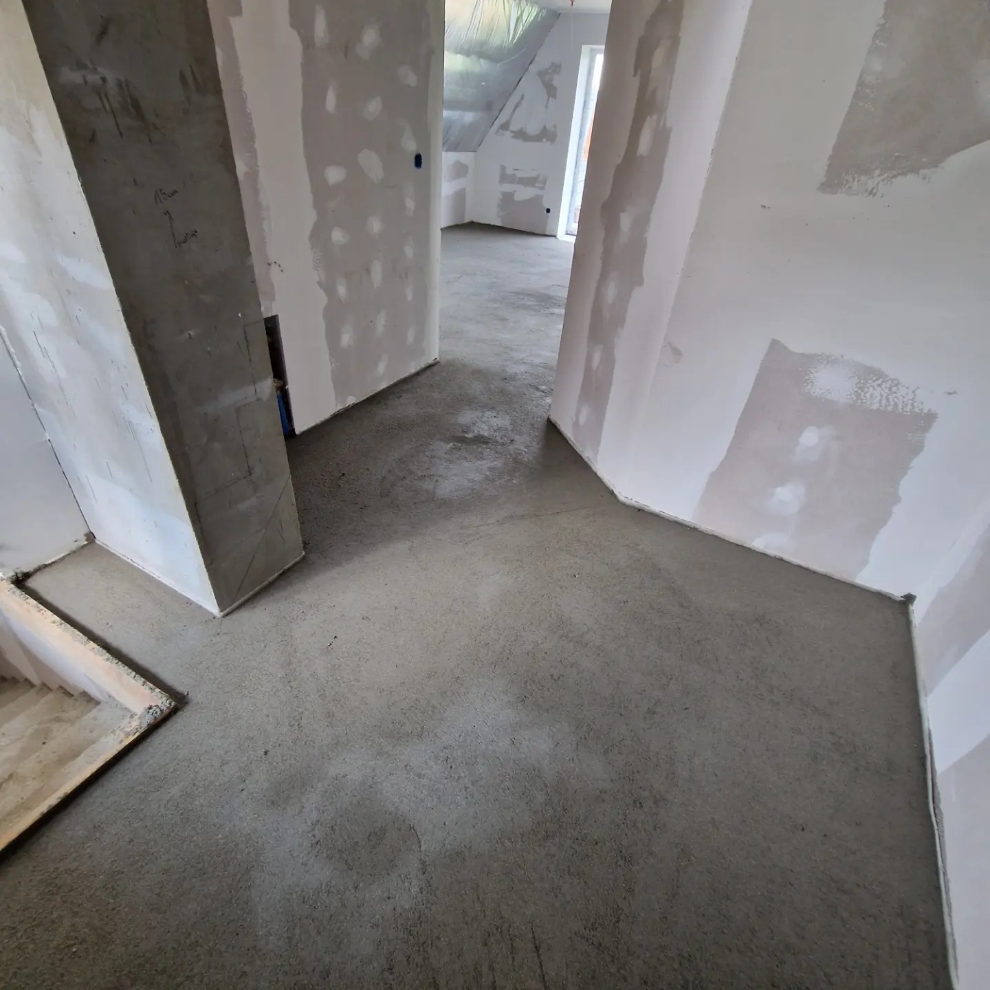 Surowy betonowy wylewka podłogowa wewnątrz budynku w trakcie prac wykończeniowych, widoczne ściany z płyt gipsowo-kartonowych przed malowaniem, schody w lewym dolnym rogu.