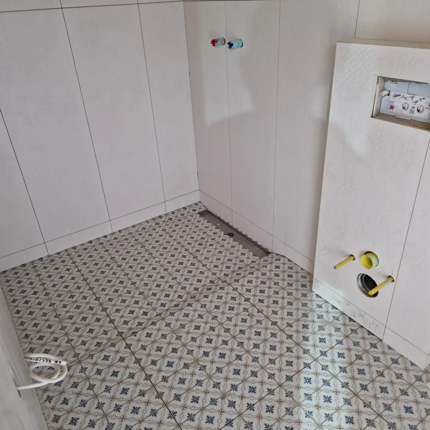 Wnętrze łazienki w trakcie remontu: białe płytki ścienne, wzorzysta podłoga z odpływem liniowym, przygotowane przyłącza wodne i stelaż podtynkowy WC.