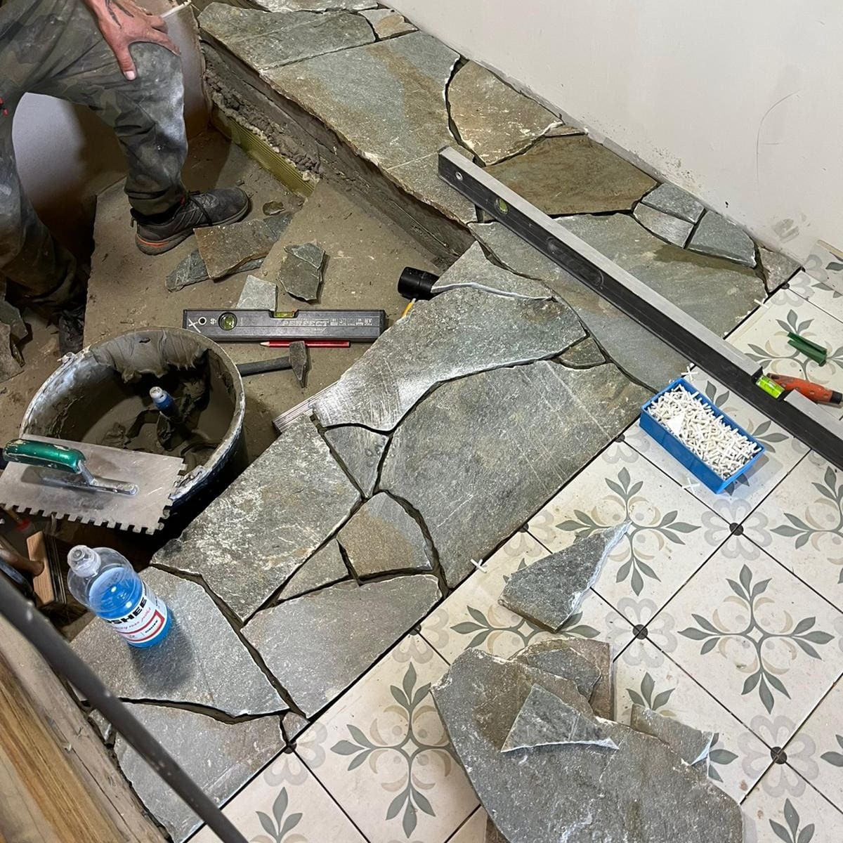 Układanie nieregularnych płyt kamiennych na podłodze, widoczne narzędzia glazurnicze, poziomica, wiadro z zaprawą, fragment ułożonej podłogi z wzorem geometrycznym.