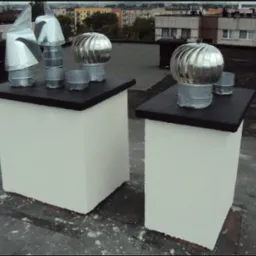 Dwa odnowione kominy na dachu bloku, z różnymi metalowymi nasadami kominowymi, w tle widoczny malarz przy pracy.