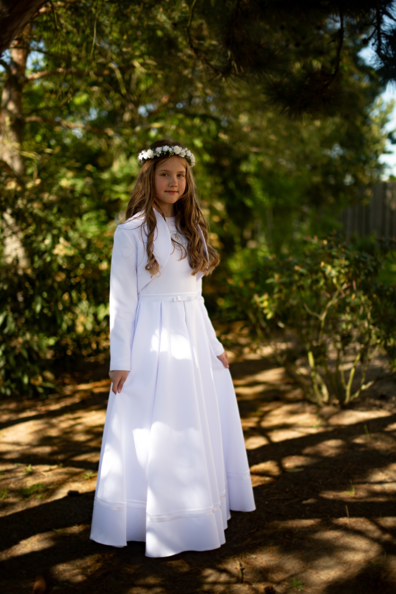 Dziewczynka w białej sukience komunijnej i wianku z białych kwiatów pozuje w ogrodzie w słoneczny dzień.