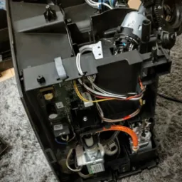 Wnętrze rozmontowanego ekspresu do kawy z widocznymi przewodami, elektroniką i elementami mechanicznymi na szarym dywanie.