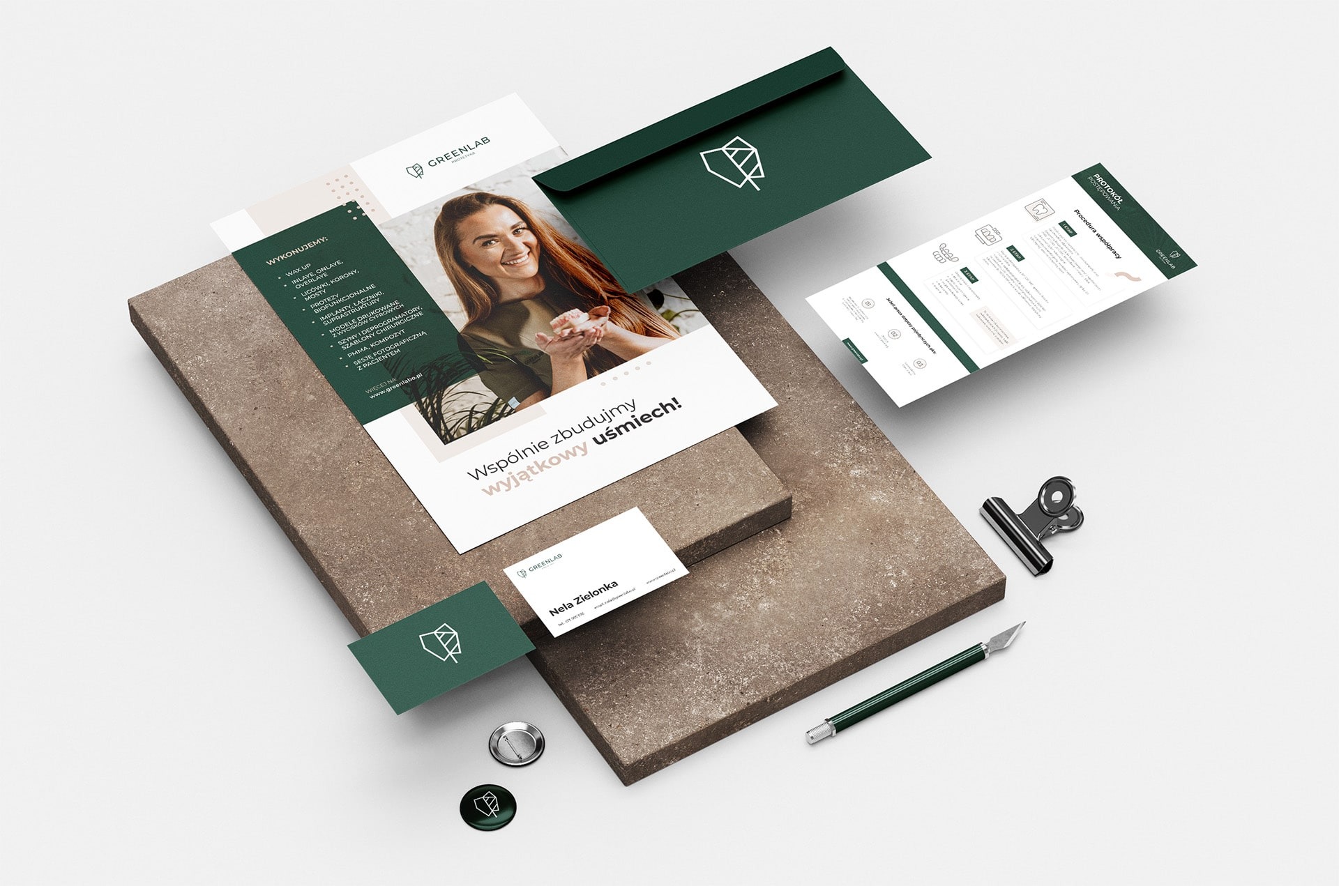 Zestaw materiałów firmowych Greenlab Protetyka: ulotka z uśmiechniętą kobietą, wizytówka, koperta, przypinki i dokument z procedurą współpracy, ułożone na szarym tle.