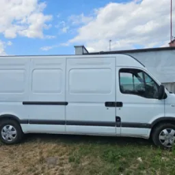 Biały samochód dostawczy Renault Master na tle betonowego muru i błękitnego nieba z chmurami.