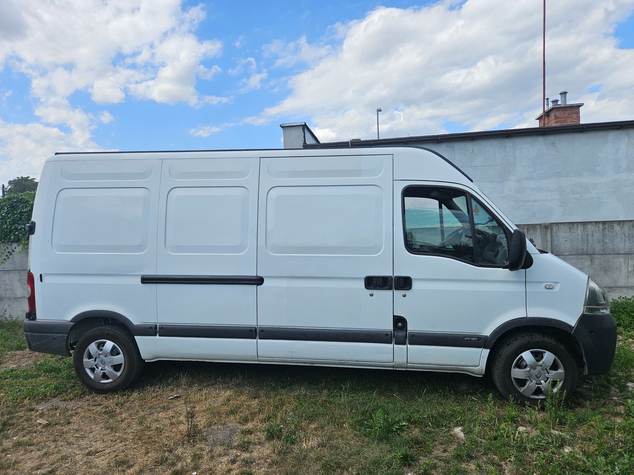 Biały samochód dostawczy Renault Master na tle betonowego muru i błękitnego nieba z chmurami.