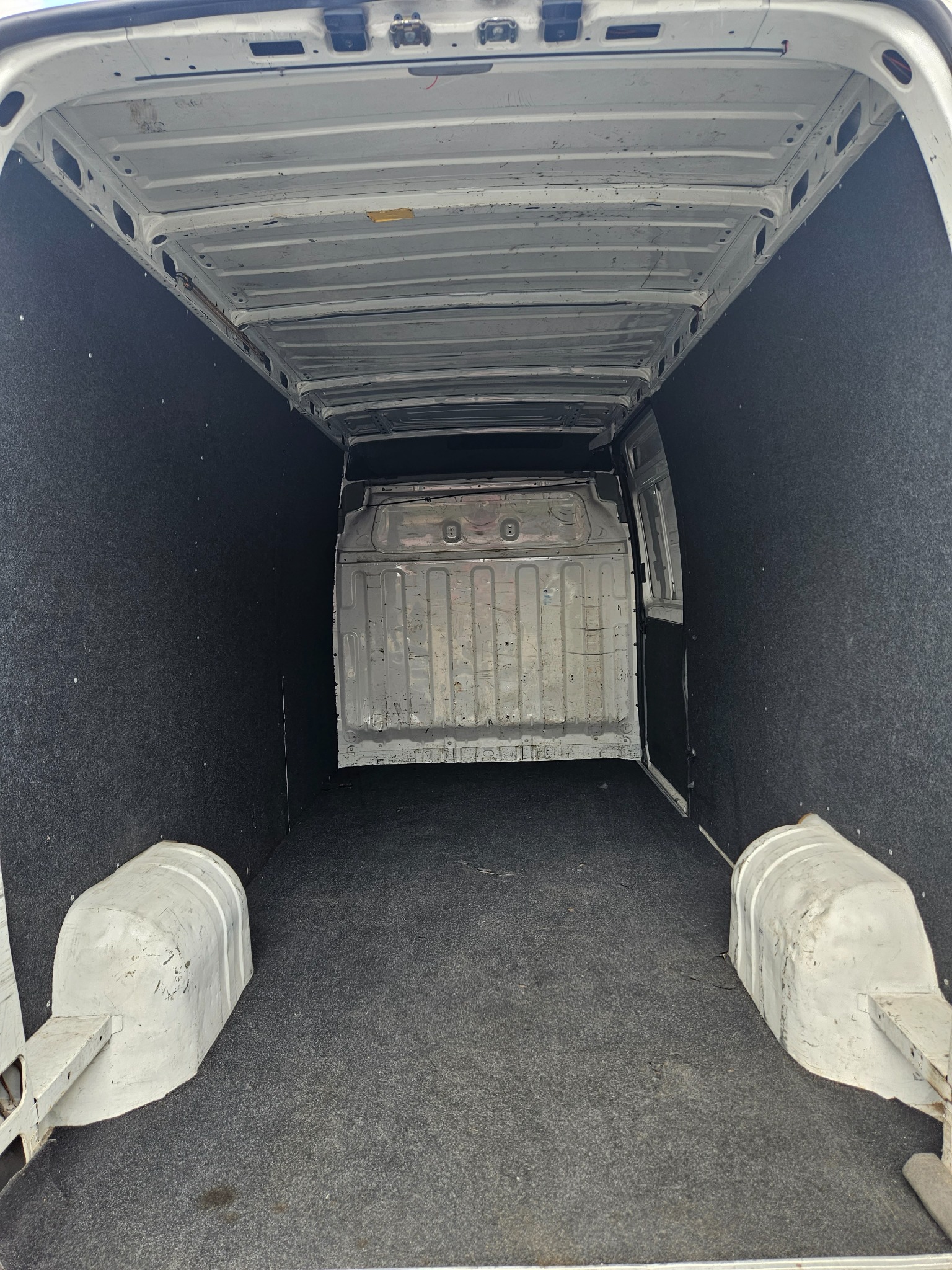 Pusta przestrzeń ładunkowa białego busa wyłożona szarą wykładziną na podłodze i czarną na ścianach, widok od tyłu pojazdu.