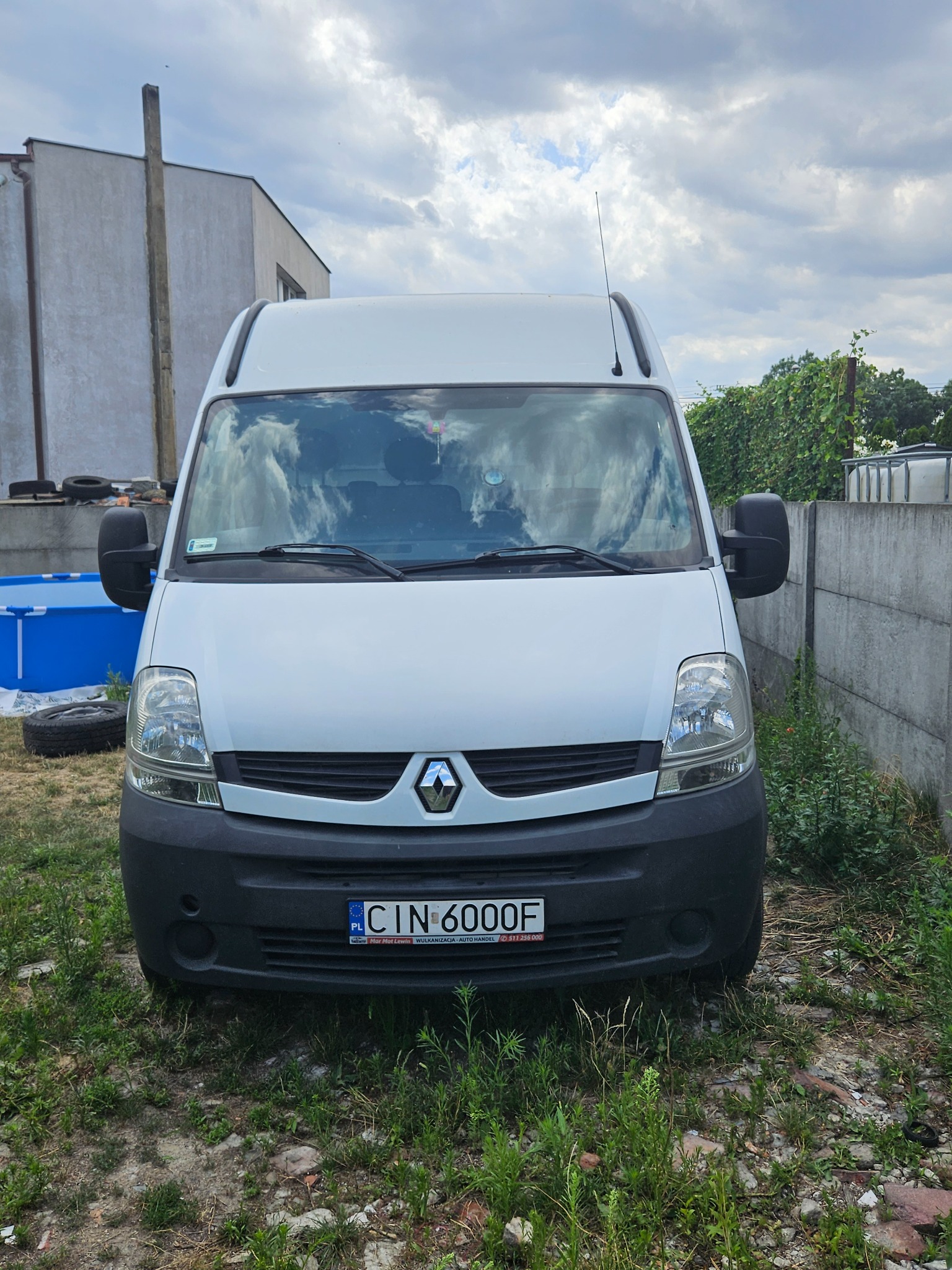 Biały bus Renault Master zaparkowany na trawiastym terenie z widocznym basenem i betonowym ogrodzeniem w tle, tablica rejestracyjna CIN 6000F.