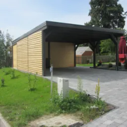 Nowoczesna wiata garażowa z jasnymi drewnianymi panelami i ciemną konstrukcją, przylegająca do budynku, z podjazdem z kostki brukowej i zadbanym trawnikiem z młodymi krzewami.