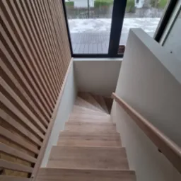 Nowoczesne schody drewniane z jasnego drewna, z ukośną ścianą z drewnianych listew i oknem dachowym z widokiem na zimowy krajobraz.