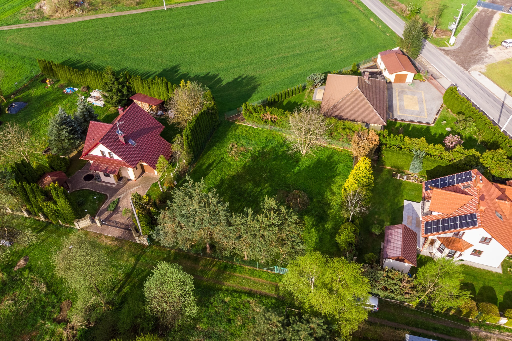 Widok z lotu ptaka na zadbany dom jednorodzinny z czerwonym dachem i panelami słonecznymi, otoczony zielonym ogrodem z wysokimi tujami i trawnikiem, sąsiadujący z polami i drogą.