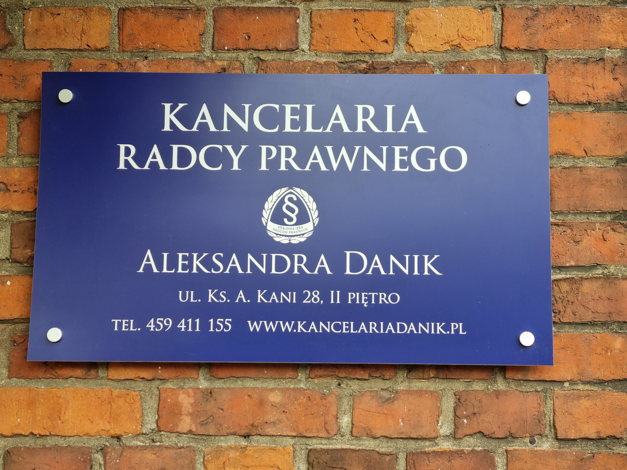 Niebieska tablica informacyjna 'Kancelaria Radcy Prawnego Aleksandra Danik' z adresem i numerem telefonu, zamocowana na ceglanej ścianie.