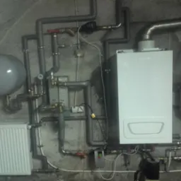 Biały piec centralnego ogrzewania z szarymi rurami, grzejnikiem i zbiornikami wyrównawczymi na tle surowej, betonowej ściany w piwnicy.