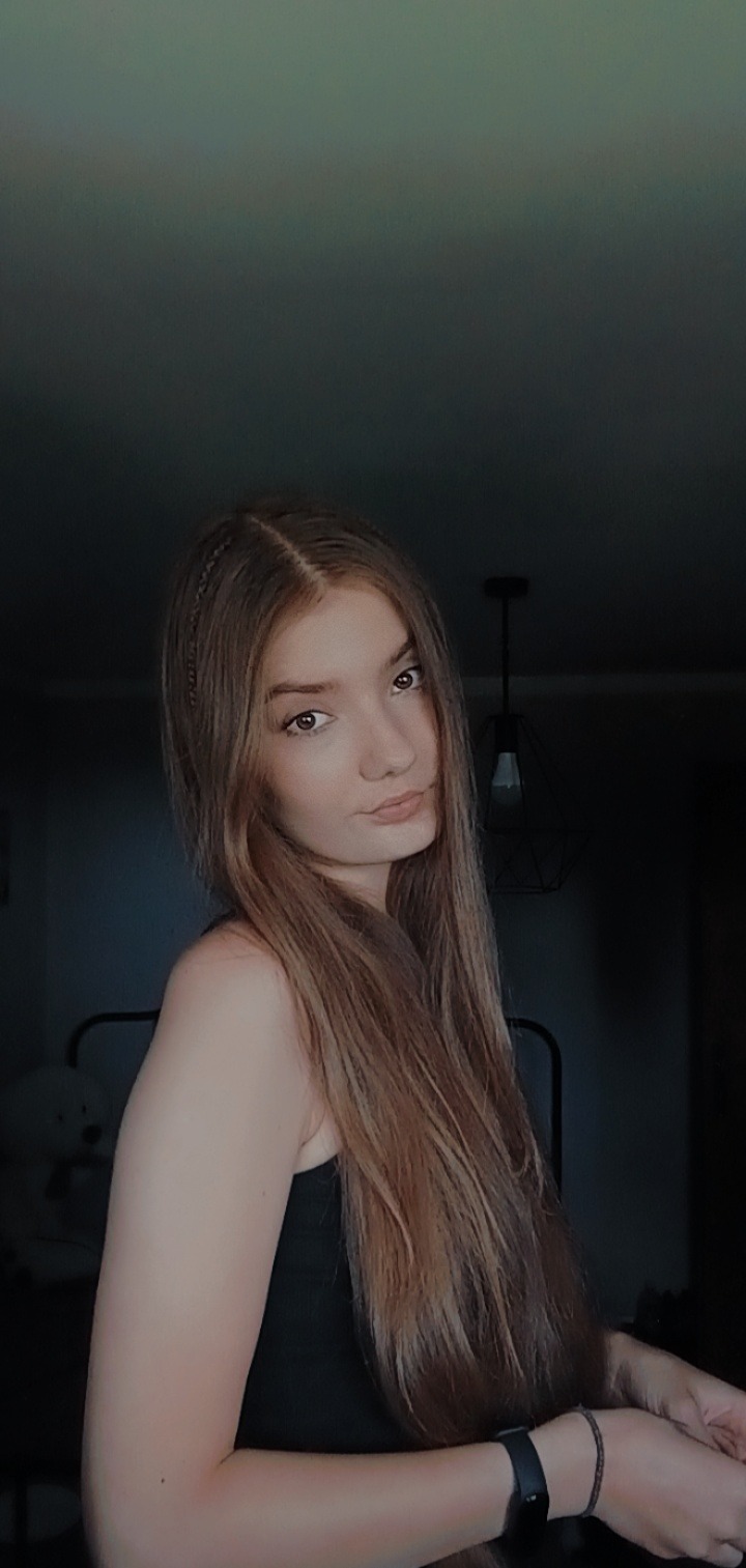 Portret młodej kobiety z długimi brązowymi włosami z wplecionymi warkoczykami, pozującej w słabym oświetleniu wnętrza z widoczną lampą i fragmentem łóżka.