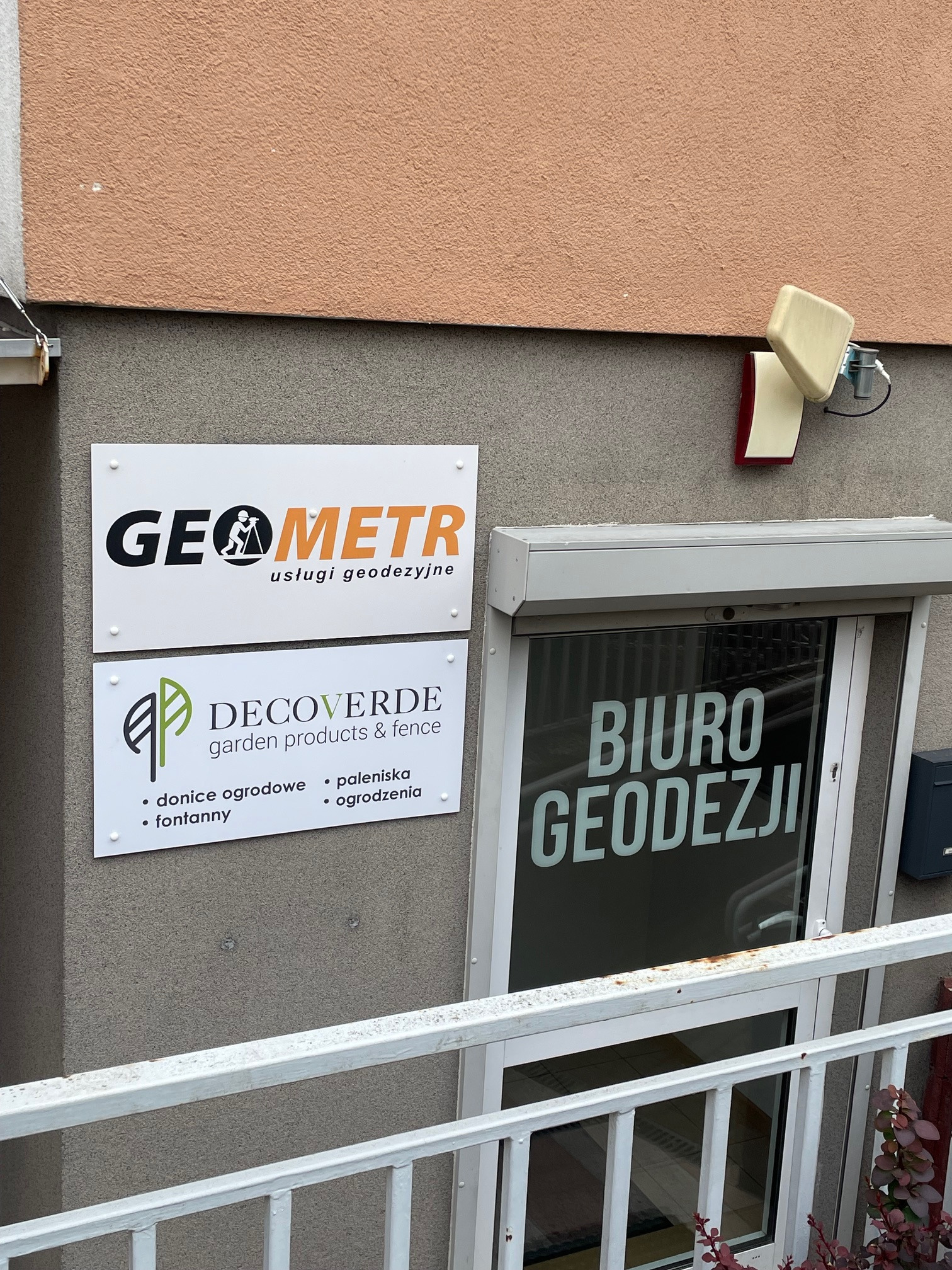 Zewnętrzny widok biura z szyldami: „GEOMETR usługi geodezyjne” oraz „DECOVERDE garden products & fence”. Na szybie okna napis: „BIURO GEODEZJI”.
