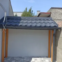 Zadaszenie nad wejściem z czarną blachodachówką i drewnianymi słupami nośnymi, widoczny fragment elewacji budynku z rurą wentylacyjną, kostka brukowa na podłożu.