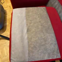 Czerwona kanapa z widoczną r&oacute;żnicą w czystości tapicerki po częściowym czyszczeniu, z lewej strony jaśniejsza i czysta, z prawej brudna, obok widoczny odkurzacz piorący.