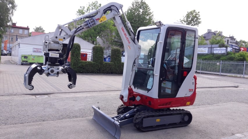 Mała maszyna budowlana Takeuchi TB216 z zamontowanym chwytakiem hydraulicznym do drewna lub kamieni, stojąca na placu w słoneczny dzień.