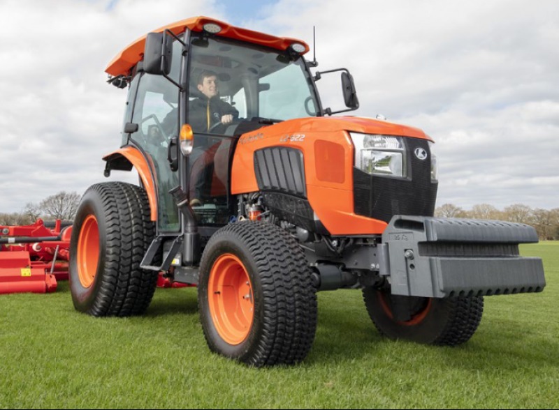 Pomarańczowy traktor Kubota z kabiną, widok z przodu na zielonym trawniku, kierowca w środku, dodatkowy osprzęt z tyłu.