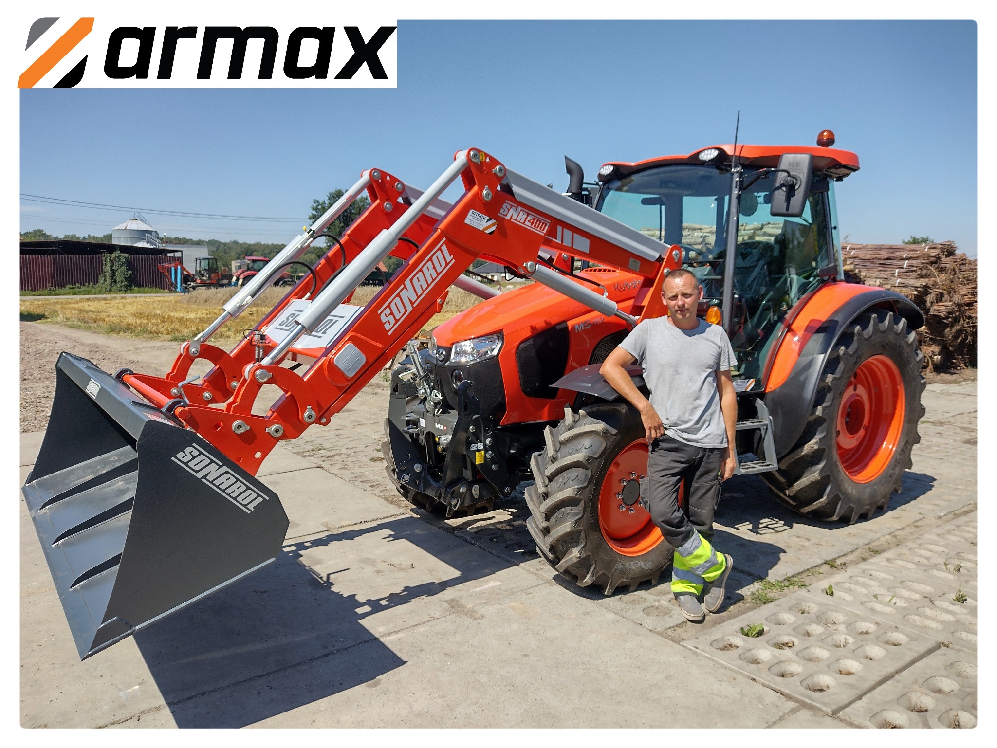 Pomarańczowy ciągnik Kubota z ładowaczem czołowym Sonarol, prezentowany przez mężczyznę w szarym T-shircie i spodniach roboczych z odblaskami, na tle wiejskiego krajobrazu.
