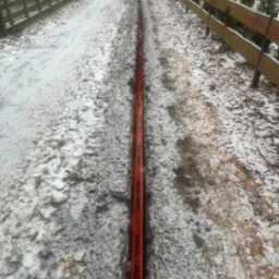 Czerwony kabel telekomunikacyjny ułożony wzdłuż drogi gruntowej pokrytej częściowo śniegiem, otoczony drewnianymi płotami.