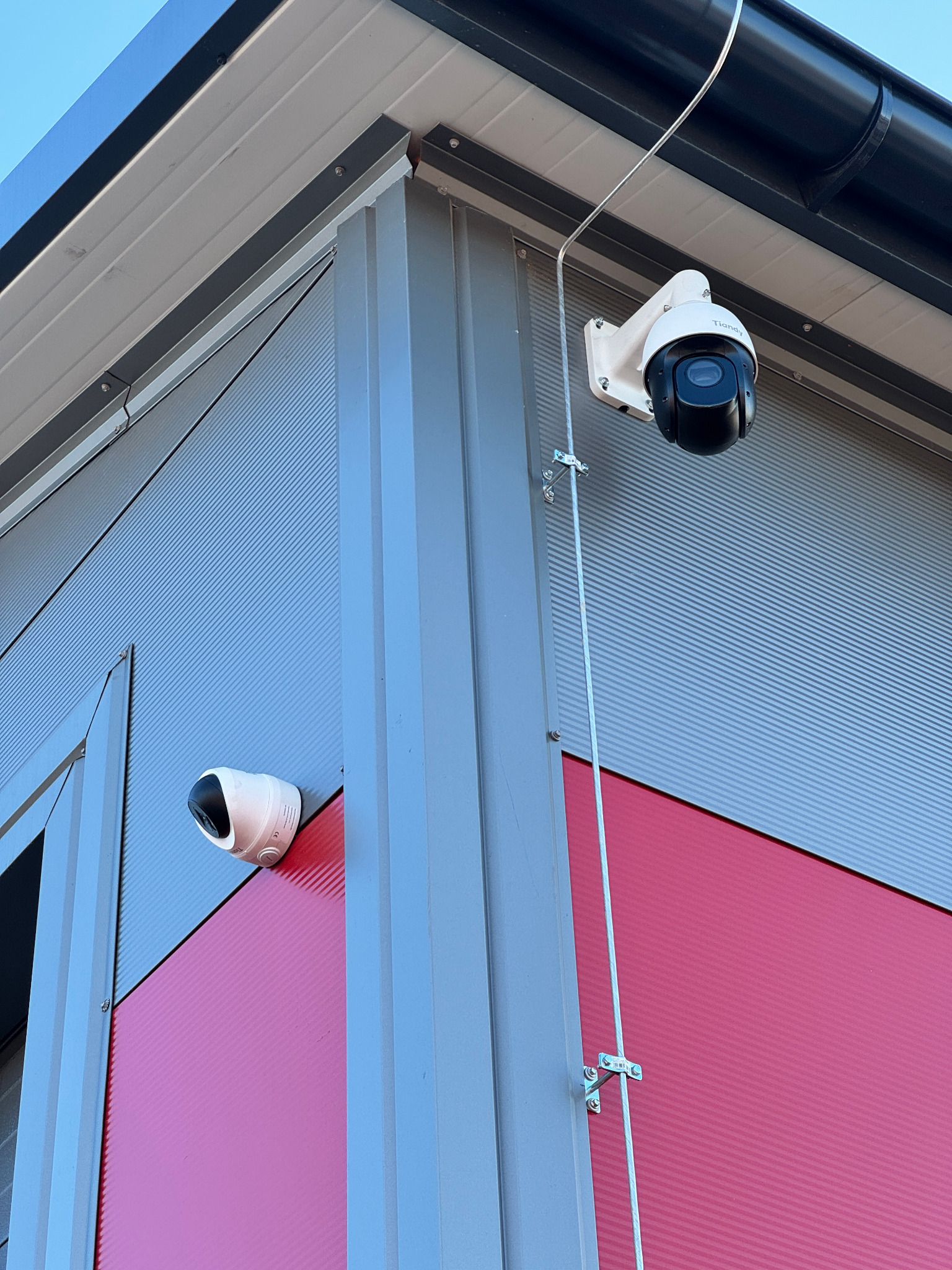 Dwie kamery monitoringu zamontowane na elewacji budynku z blachy trapezowej w kolorach szarym i różowym. Górna kamera PTZ z kablem w metalowej listwie. Dolna kamera kopułkowa.