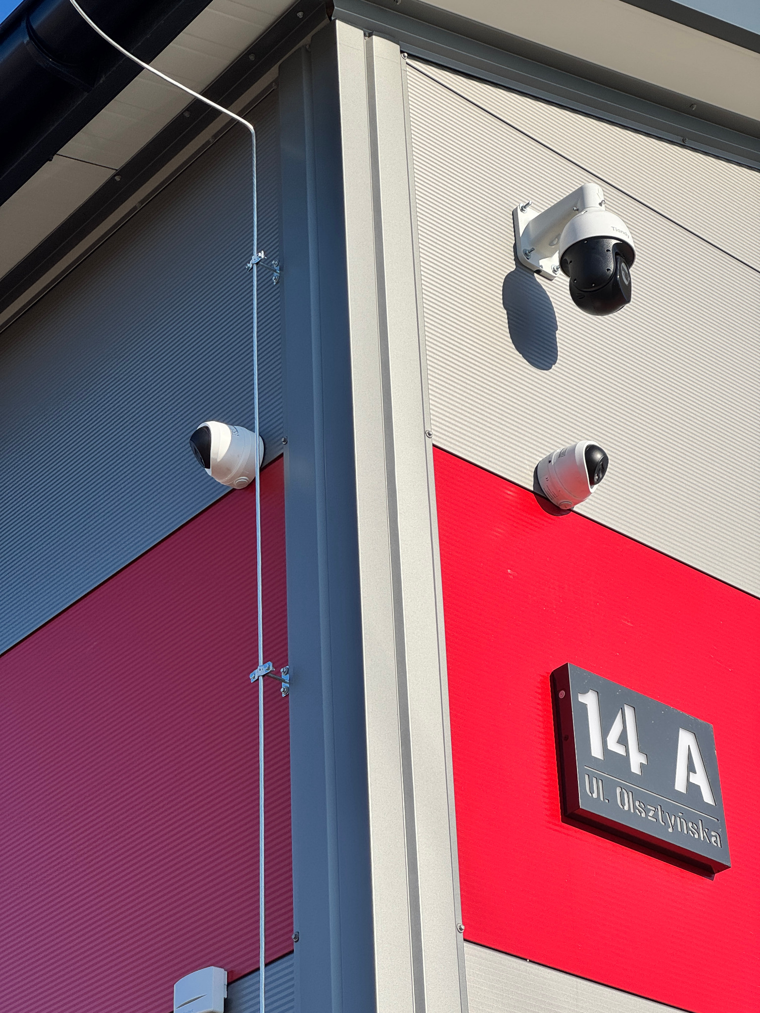 Dwie kamery kopułkowe i jedna obrotowa PTZ zamontowane na elewacji budynku o szaro-czerwonej kolorystyce, widoczny adres budynku na tabliczce.