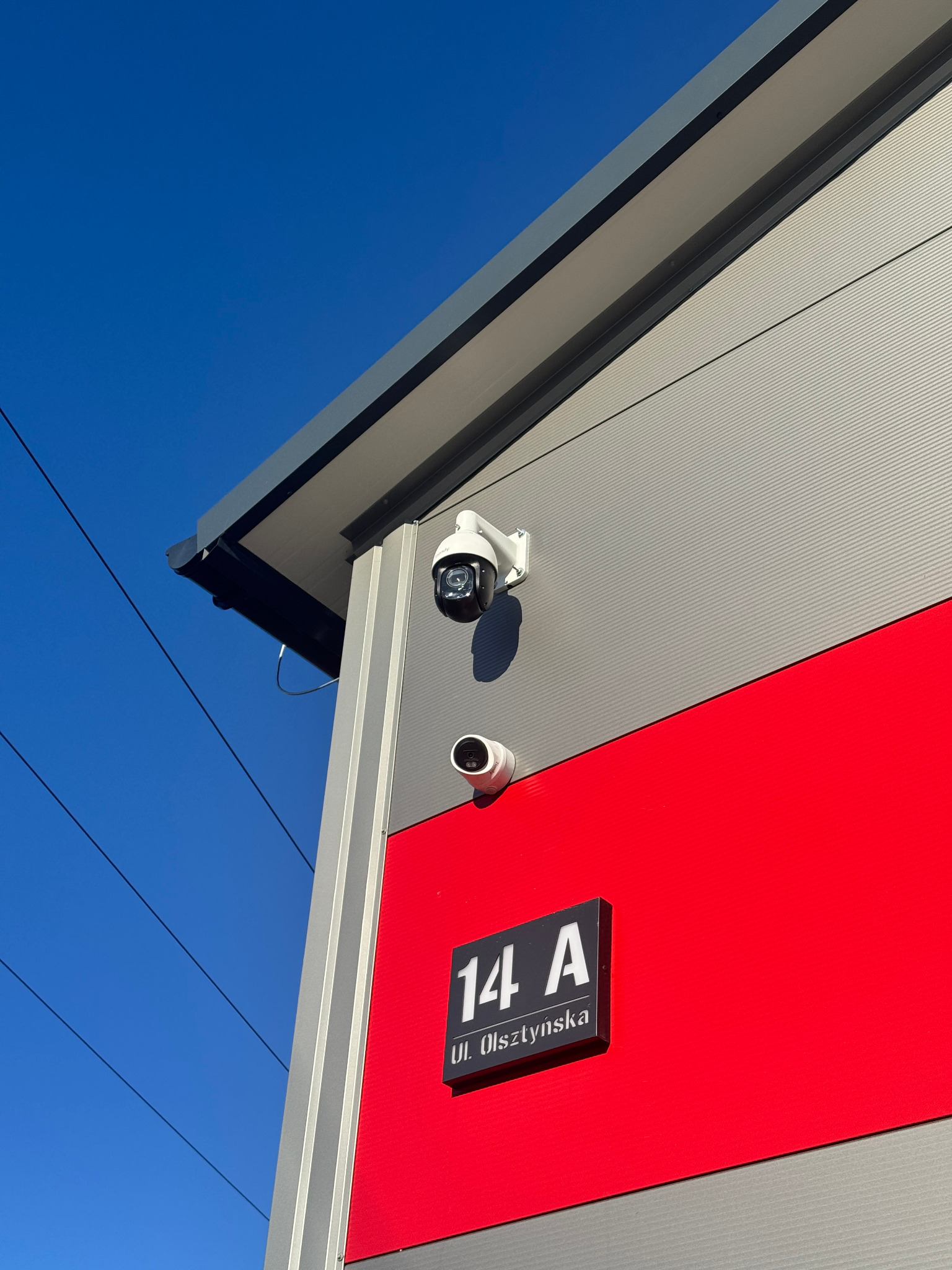 Dwie kamery monitoringu zamontowane na elewacji budynku z numerem adresowym 14A Ul. Olsztyńska, na tle błękitnego nieba.