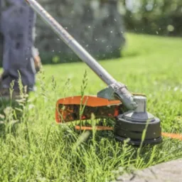 Przycinanie trawy przy krawężniku za pomocą podkaszarki spalinowej; w powietrzu unoszą się drobne źdźbła trawy.