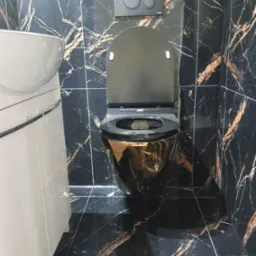 Toaleta wisząca w kolorze czarnym ze złotymi refleksami, zamontowana na ścianie z czarnych płytek imitujących marmur ze złotymi żyłami, obok biała umywalka z szafką.