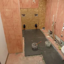 Układanie ciemnych płytek podłogowych w łazience, widoczne poziomice, wiadra z zaprawą i system poziomowania płytek, ściany częściowo wyłożone jasnymi płytkami, fragmenty ścian z płyt OSB.