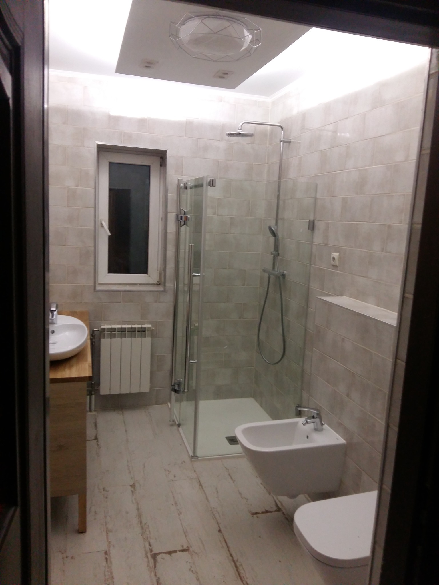 Wykończona łazienka z kabiną prysznicową, umywalką nablatową na drewnianej szafce, bidetem i grzejnikiem żeberkowym pod oknem, wykończona jasnymi płytkami ceramicznymi.