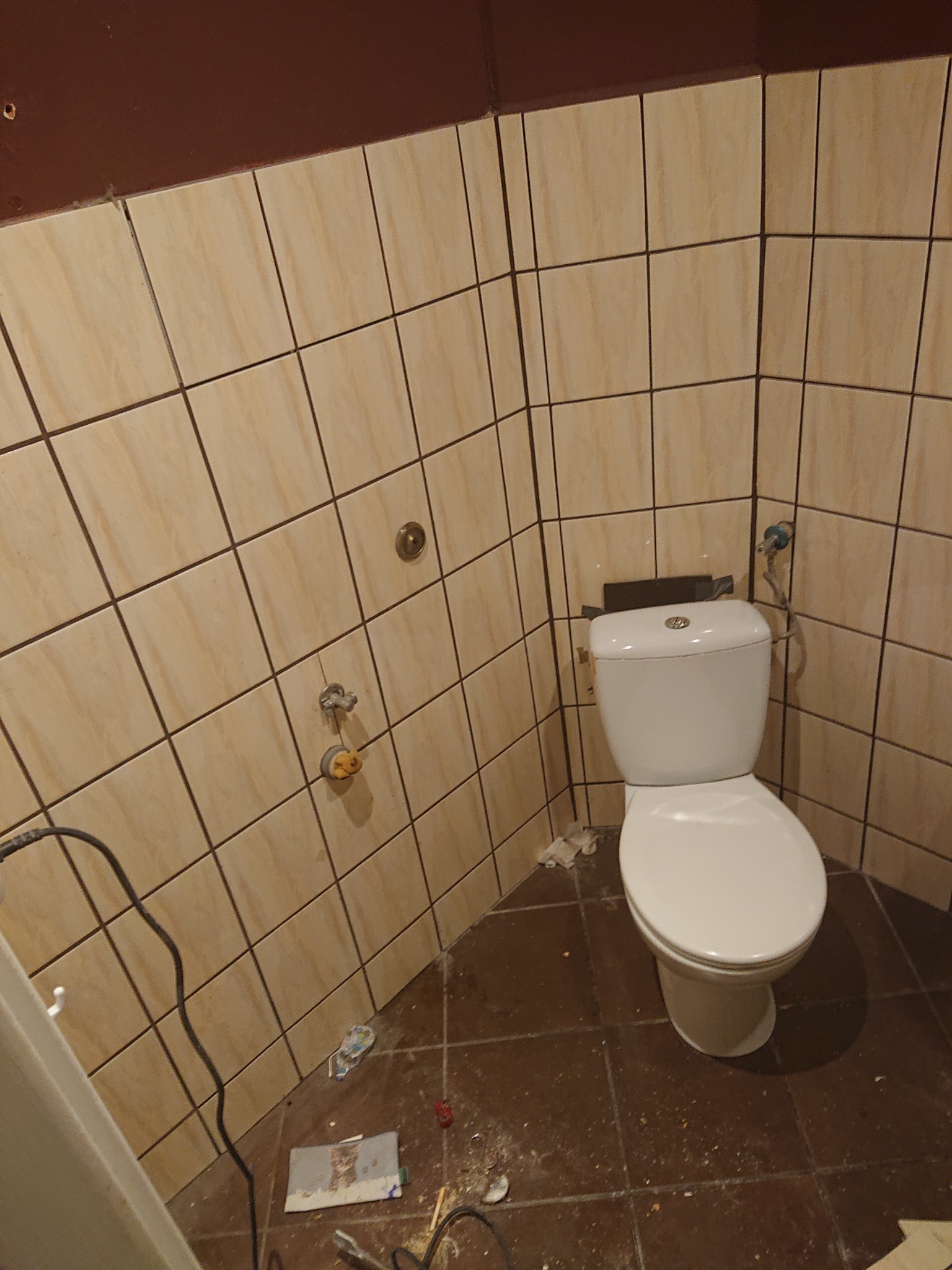 Biała toaleta w trakcie montażu w łazience z beżowymi płytkami na ścianach i brązowymi na podłodze, widoczne przewody i drobne śmieci remontowe.